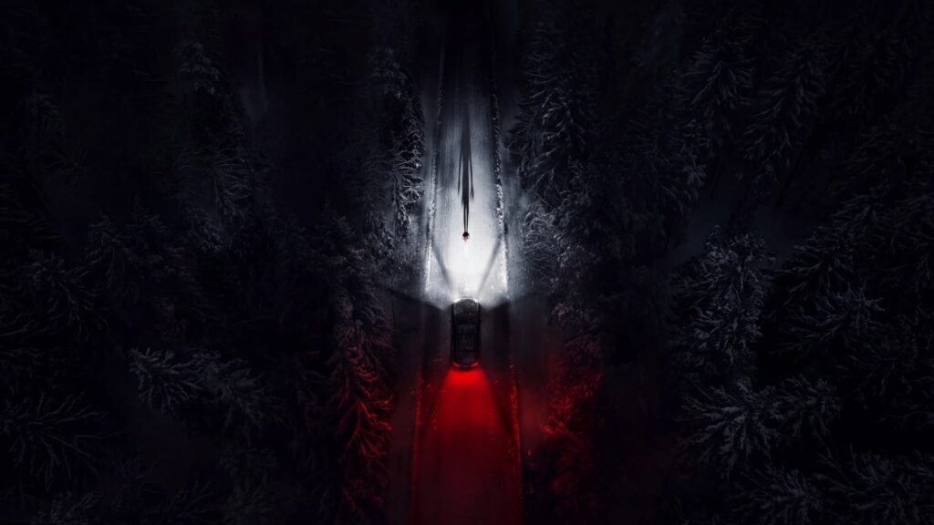 Top-down nighttime view of a car on a narrow snow-lined forest road, its red rear lights glowing beneath a small lone figure in a bright patch of snow.