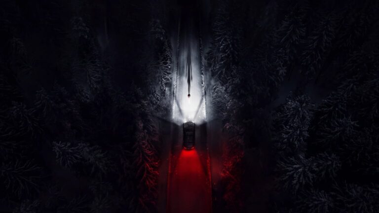 Top-down nighttime view of a car on a narrow snow-lined forest road, its red rear lights glowing beneath a small lone figure in a bright patch of snow.