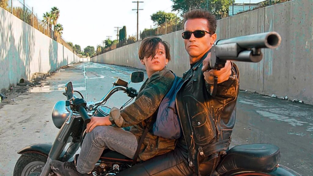 A leather-jacketed man in dark sunglasses rides behind a teenage boy on a motorcycle in a concrete flood channel, aiming a shotgun toward the viewer.