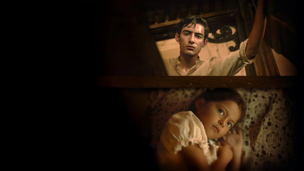 Dark, sepia-toned cinematic scene of a worried young man leaning over a wooden rail above a child lying on patterned bedding, with heavy shadows and empty black space on the left.