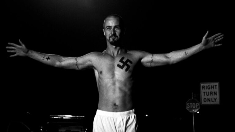 Black-and-white still of a shirtless bald man with arms outstretched, showing a large swastika tattoo on his chest, beside stop and “RIGHT TURN ONLY” road signs at night.