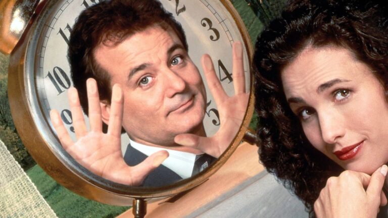 Two people pose beside a large clock, with a man raising both hands in front of the clock face and a woman looking toward the camera.