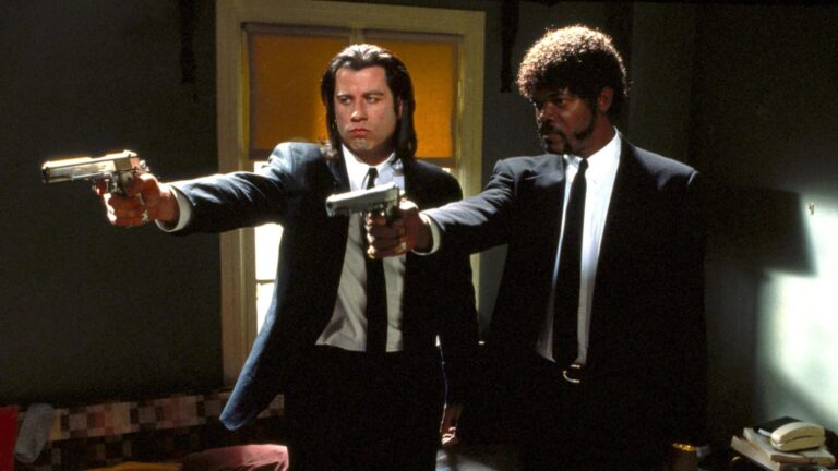 Cinematic still of two men in black suits and ties standing in a dim room, each aiming a handgun toward the left.