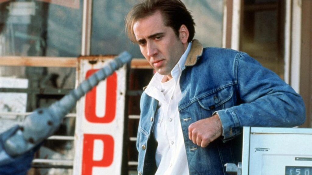 A man in a blue denim jacket leans beside a gas pump, looking downward, with a blurred motorcycle handlebar in the foreground and large partial red lettering behind him.