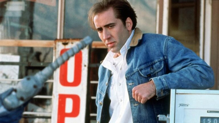 A man in a blue denim jacket leans beside a gas pump, looking downward, with a blurred motorcycle handlebar in the foreground and large partial red lettering behind him.
