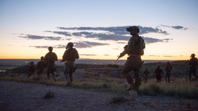 Cinematic widescreen scene of silhouetted armed tactical personnel walking across scrubland at sunset beneath a pale blue sky with scattered clouds.