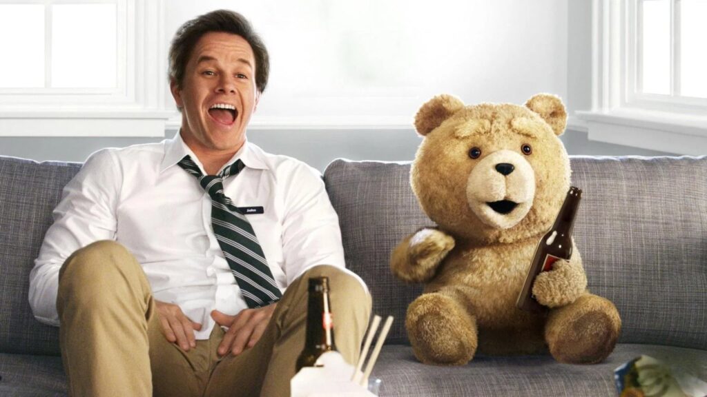 A smiling man in a loosened tie sits on a gray sofa beside a large tan teddy bear holding a brown beer bottle; another bottle and takeout containers sit in the foreground.