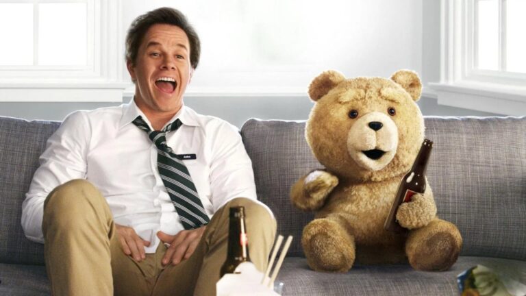 A smiling man in a loosened tie sits on a gray sofa beside a large tan teddy bear holding a brown beer bottle; another bottle and takeout containers sit in the foreground.