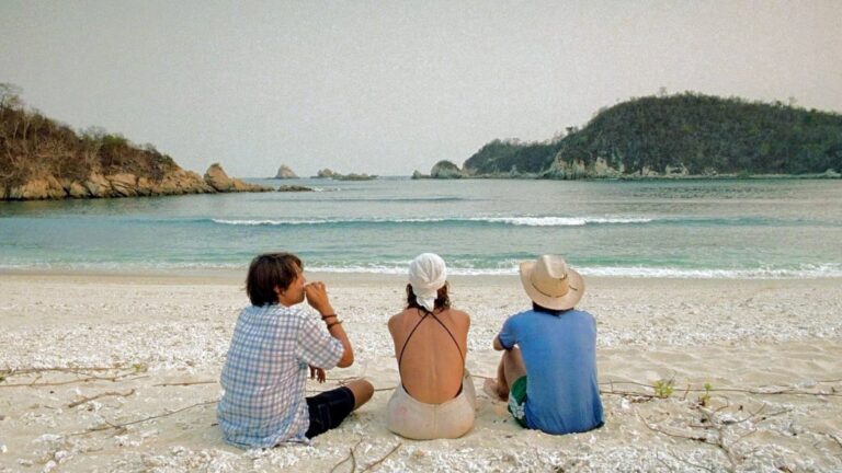 Three people sit with their backs to the camera on a pale sandy beach, facing calm blue water and forested rocky headlands under a hazy sky.